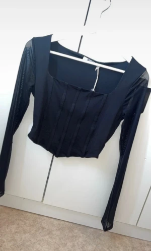 Svart mesh korsettopp från NA-KD - Säljer en svart långärmad topp från NA-KD med mesh-ärmar. Toppen har fyrkantig ringning och tight passform som framhäver siluetten. Perfekt för dig som vill ha en edgy och trendig look.