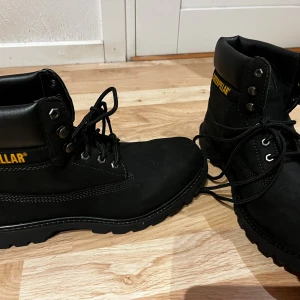 Svarta boots från Caterpillar oanvända - Stilrena svarta boots från Caterpillar med grov sula och snörning. Skorna har rund tå, robust design och detaljer i gult på sidan och plösen. Tillverkade i slitstark mocka och skinn, perfekta för dig som gillar en tuff look. Säljes pga felköp