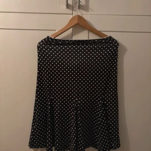 Svart prickig kjol med volangkant - Svart kjol med vita prickar och volangkant nedtill. Kjolen har en figurnära passform upptill och blir lite vidare mot knäna. Materialet är mjukt och stretchigt, perfekt för dig som gillar retrovibbar och prickigt mönster. Söt spetsdetalj vid midjan.Polka dot midi/knähöjd kjol, strechig material, volanger på änden. Helt ny med prislapp på! Sista bilderna vissar hur den sitter på