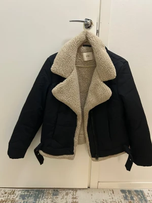 Svart teddyjacka med beige foder från STR - Svart teddyjacka med fluffigt beige foder och bred krage. Jackan har dragkedja framtill och coola bältesdetaljer vid nederkanten. Perfekt för dig som vill ha en varm och snygg look under höst och vinter.