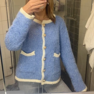 Blå kofta med vita detaljer - Mysig blå kofta med vita kanter och knappar framtill. Koftan har två fickor och är stickad i ett fluffigt material som känns skönt mot huden. Perfekt för kyliga dagar och har långärmad passform med normal passning.