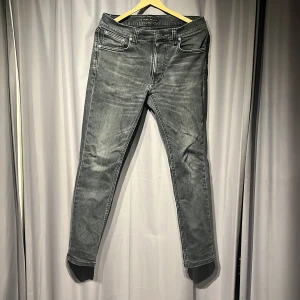 Grå jeans från Nudie Jeans Co - Snygga grå jeans från Nudie Jeans Co med klassisk femficksdesign och svarta broderade detaljer på bakfickorna. Jeansen har en rak passform och är tillverkade i slitstarkt denim med cool tvättad look. Nypris 1600kr