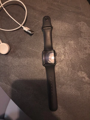 Apple Watch Series 3 38mm svart - Apple Watch Series 3 med 38mm aluminiumboett i svart och matchande svart silikonarmband. Klockan har en stilren, fyrkantig design med digital krona och pulsmätare på baksidan. Perfekt för dig som gillar teknik och vill ha koll på din vardag direkt på handleden.