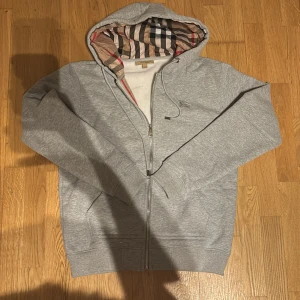 Grå hoodie från Burberry med dragkedja - Snygg grå hoodie från Burberry med klassiskt rutigt foder i huvan och insidan. Tröjan har dragkedja framtill, snörning i huvan och två fickor. Tillverkad i mjuk bomullsmix som känns riktigt skön mot huden. Perfekt för dig som vill ha en stilren men lyxig look.