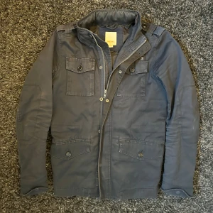 Mörkblå field jacket från Diesel - Snygg mörkblå parkajacka från Diesel med flera fickor framtill, både med knapp och dragkedja. Jackan har hög krage, axelklaffar och coola detaljer som ger en urban vibe. Perfekt för dig som gillar streetstyle och funktionella plagg.