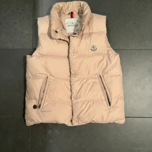 Beige dunväst från Moncler - Snygg beige dunväst från Moncler med hög krage och ikonisk logga på bröstet. Västen har två fickor med dragkedja och tryckknappar framtill. Perfekt för lager-på-lager och har en clean, puffad look som är både trendig och bekväm. Priset kan diskuteras 