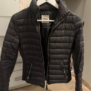Svart quiltad pufferjacka från Zara - Svart pufferjacka från Zara med quiltad design och hög krage. Jackan har dragkedja framtill, två fickor med dragkedja och snygga detaljer på ryggen. Materialet är syntet och jackan har en slim passform som sitter snyggt på kroppen.