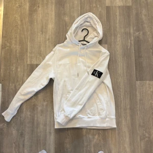 Vit hoodie från Calvin Klein Jeans - Vit hoodie från Calvin Klein Jeans med svart CK-logga på ärmen. Tröjan har huva med snörning, magficka och ribbade muddar vid ärmslut och nederkant. Perfekt för dig som gillar clean och stilren streetwear. Tröjan är i storlek S men passar även Xs. Nypris i affär är ca 700-800 kr, mitt pris går att diskutera.