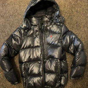 Svart pufferjacka från Polo Ralph Lauren - Svart pufferjacka från Polo Ralph Lauren med glansig finish och huva. Jackan har dragkedja framtill, två sidofickor med dragkedja och ett rött Polo-logomärke på bröstet. Kvitto finns.