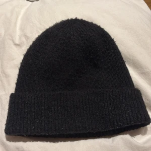 Svart ribbad mössa från KappAhl - Svart mössa från KappAhl i ribbstickad design. Klassisk form med uppvikt kant, perfekt för kalla dagar. Materialet är mjukt och stickat, vilket gör den skön att bära. Enkel och stilren look som funkar till de flesta outfits.