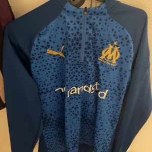 Marseille fotbollströja - Säljer en blå långärmad fotbollströja från Puma för Olympique de Marseille. Tröjan har ett geometriskt mönster i olika blå nyanser, halv dragkedja och klubbmärke samt Puma-logga i gult på bröstet. Perfekt för dig som älskar fotboll och snygg sportstil.