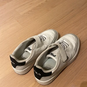 Vita sneakers från Fila med kardborre - Snygga vita Fila sneakers med svarta detaljer på hälen och grå mönstrade partier. Skorna har både snörning och kardborreband för enkel användning. Ovandelen är i syntetmaterial och sidorna har perforeringar för extra ventilation. Perfekt för dig som gillar sportig stil.