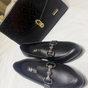 Svarta loafers med glittrig detalj - Snygga svarta loafers från Moment Shoes med spetsig tå och glittrig metallkedja över foten. Skorna är tillverkade i skinnimitation och har en platt sula för bekväm användning. Perfekt för dig som gillar stilrena och trendiga skor med en extra touch.             Pris för både 350.   Frakten står ni för