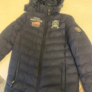 Mörkblå pufferjacka från Vinson Polo Club - Snygg mörkblå pufferjacka från Vinson Polo Club med broderade loggor på bröstet och ärmen. Jackan har huva, dragkedja framtill och två sidofickor med dragkedja. Perfekt för kalla dagar och har en sportig vibe med coola detaljer.                   Köparen står för frakten.