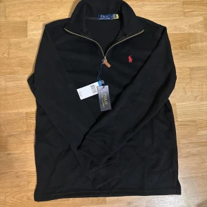 Svart half zip tröja Polo Ralph Lauren - Svart långärmad tröja från Polo Ralph Lauren med half zip och klassisk röd broderad logga på bröstet. Tröjan har en stilren ståkrage och är gjord i mjuk bomull, perfekt för dig som gillar clean och enkel stil.