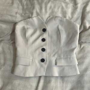 Beige tubtopp med knappar från new yorker - Säljer en stilren beige tubtopp med svarta knappar framtill. Toppen har en skräddad look med markerade sömmar och dekorativa fickdetaljer. Perfekt för dig som vill ha en chic och unik topp till din outfit.