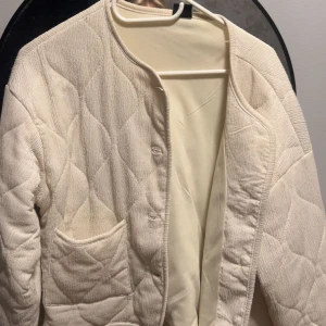 Beige quiltad cardigan jacka - Quiltad cardigan jacka i beige/vit med snyggt mönstrad yta och rund hals. Jackan har en praktisk ficka framtill och knäppning längs framsidan. Materialet är mjukt och har en lätt struktur, perfekt för lager-på-lager under höst och vinter.