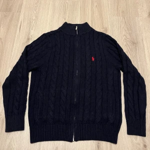 Mörkblå kabelstickad kofta Polo Ralph Lauren - Snygg mörkblå kabelstickad kofta från Polo Ralph Lauren med röd broderad logga på bröstet. Koftan har ribbad krage, muddar och nederkant samt dragkedja framtill. Perfekt för dig som gillar klassisk stil med en modern touch.