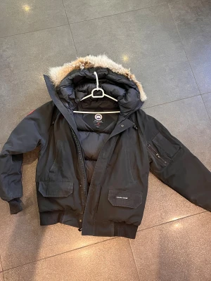 Svart dunjacka Canada Goose med päls - Svart dunjacka från Canada Goose med fluffig päls på huvan. Jackan har flera fickor, ribbade muddar och dragkedja framtill. Perfekt för kalla vinterdagar och har en klassisk, urban look. Märket syns tydligt på axeln och insidan.