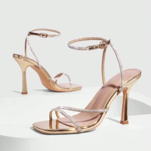 Glittriga guldiga klackar - Säljer ett par sandaler med hög klack (ca 10 cm) i metallic guld. Skorna har smala glittriga remmar med strassdetaljer, fyrkantig tå och justerbart spänne runt ankeln. Insidan är ljusbeige och sulan har en lyxig finish. Perfekta för att sticka ut och ge extra bling till din outfit.