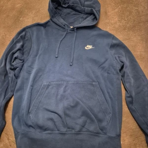 Blå hoodie från Nike med huva - Säljer en blå hoodie från Nike med klassisk logga på bröstet. Tröjan har huva med snörning och en stor magficka framtill. 
