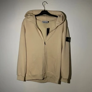 Stone Island Hoodie - Snygg beige hoodie från Stone Island med klassisk logotyp-patch på vänster ärm. Tröjan har huva, dragkedja framtill och två fickor. Tillverkad i mjuk bomull och har en relaxed passform med långa ärmar. Tröjan är i Storlek L, perfekt inför vintern!🙌