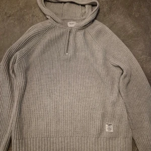 Grå stickad hoodie från Pepe Jeans - Säljer en grå stickad hoodie från Pepe Jeans med ribbad struktur och half zip framtill. Tröjan har huva och långa ärmar.. Märkeslapp nedtill på framsidan.
