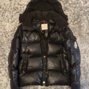 Svart Moncler pufferjacka med huva - Svart pufferjacka från Moncler med glansig yta, avtagbar huva och två dragkedjeförsedda fickor fram. Jackan har klassisk Moncler-logga på ärmen och quiltad design som ger en schysst volym. Perfekt för kalla dagar när du vill hålla stilen.