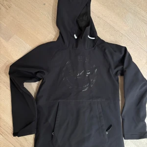 Svart skidjacka från 8848 Altitude - Svart vindjacka från 8848 Altitude med stor logga på bröstet. Jackan har huva med vit snörning, lång ärm och en stor ficka framtill med dragkedja. Insidan är fodrad med mjuk fleece för extra komfort. Perfekt för dig som gillar stilren och sportig look.