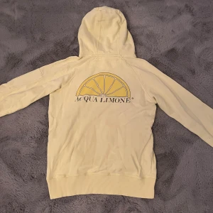 Gul hoodie Acqua Limone Lemon S - Snygg ljusgul hoodie från Acqua Limone i storlek S. Tröjan har huva med snörning, ribbade muddar och en dragkedja på ärmen. På ryggen finns ett stort citrontryck och logga, och på ärmen står märket tryckt. Perfekt för chill dagar.
