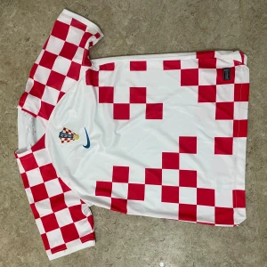 Kroatien fotbollströja Nike rutig - Säljer en officiell Kroatien fotbollströja från Nike med klassiskt rödvitt rutmönster och korta ärmar. Tröjan har landslagets emblem på bröstet och är tillverkad i lätt, ventilerande polyester. Perfekt för match eller häng med polarna.