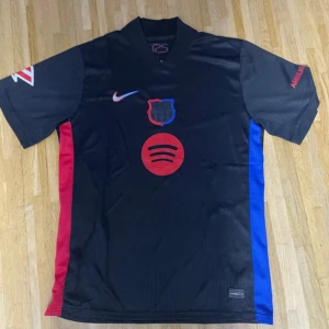 Svart FC Barcelona fotbollströja Nike - Säljer en svart FC Barcelona fotbollströja från Nike med röd och blå sidopanel, Spotify-logga på bröstet och klubbmärke i blått och rött. Tröjan har korta ärmar och är tillverkad i lätt, ventilerande polyester. UNHCR-tryck på ryggen och detaljer på ärmarna.