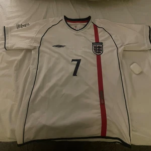 England Beckham #7 fotbollströja Umbro - Säljer en klassisk England fotbollströja från Umbro med nummer 7 och Beckham på ryggen. Tröjan är vit med svarta detaljer och en röd vertikal rand på framsidan. England-emblem på bröstet och V-ringad hals. Perfekt för dig som gillar retro och landslag.