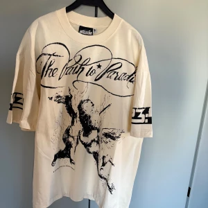 Beige t-shirt från Hellstar med tryck - Snygg oversized beige t-shirt från Hellstar med svart grafiskt tryck fram och bak. Texten 'The Path to Paradise' och coola motiv ger en unik vibe. Tryck även på ärmarna. Perfekt för dig som gillar streetwear och statement-plagg.