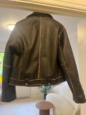 Svart bikerjacka från Zara - Snygg svart bikerjacka från Zara med silverfärgade dragkedjor och knappar. Jackan har klassisk krage, flera fickor med lock och zip, samt detaljerade sömmar och justerbara remmar vid midjan. Materialet är skinnimitation med en matt finish och långärmad design.