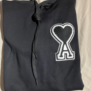 Svart hoodie från Ami med hjärtdetalj - Svart hoodie från Ami Alexandre Mattiussi med stor broderad hjärtdetalj och bokstaven A på bröstet. Tröjan har huva med snörning, ribbade muddar och magficka. Perfekt för dig som gillar stilrena streetwear-plagg med coola detaljer. Helt ny och aldrig använd då den är för liten. Den är i storlek M men skulle säga att den är S då den har lite korta armar 