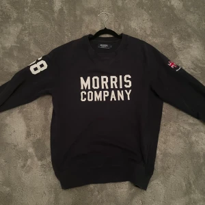 Mörkblå sweatshirt Morris Company S - Mörkblå sweatshirt från Morris Company i storlek S. Tröjan har vit text på bröstet, siffran 98 på ena ärmen och en liten brittisk flagga på andra ärmen. Rund hals och långa ärmar, tillverkad i mjuk bomull. Perfekt för dig som gillar sportig och stilren look.