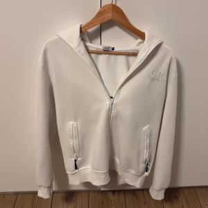 Vit hoodie med glasögon från C.P. Company - Säljer en vit hoodie från C.P. Company med ikoniska glasögon i huvan. Tröjan har dragkedja framtill, två fickor med dragkedja och broderad logga på bröstet. Mjuk insida och långärmad modell, perfekt för dig som gillar streetwear och unika detaljer. Vet ej om den är äkta då jag köpte den här på Plick, näst intill oanvänd.