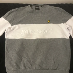 Grå och vit sweatshirt från Lyle & Scott - Snygg grå och vit sweatshirt från Lyle & Scott med broderad logga på bröstet och stor text över mitten. Tröjan har långärmade ärmar med breda vita paneler och rund hals. Mjuk bomullskvalitet och normal passform, perfekt för chill dagar.