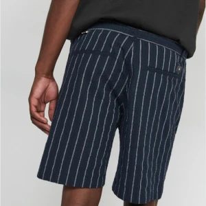 Marinblå randiga shorts från Martinique  - Snygga marinblå shorts med vita vertikala ränder från Martinique. Modellen har normal passform, midwaist och praktiska fickor både fram och bak. Perfekta för varma dagar och har en stilren look som funkar till t-shirt eller skjorta.