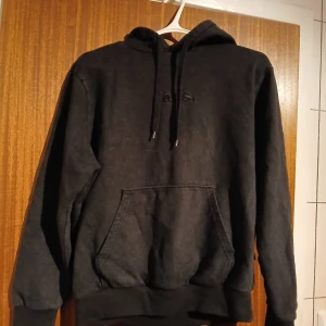 Svart hoodie från Vans med huva - Svart hoodie från Vans i relaxed fit med klassisk känguruficka och snörning i huvan. Perfekt för dig som gillar en enkel och stilren look. Mjuk insida och diskret broderad logga framtill.