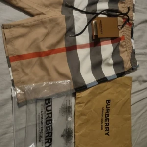 Burberry badshorts med klassiskt mönster - Säljer ett par Burberry badshorts i storlek M med ikoniskt randigt och rutig mönster i beige, vit, svart och röd. Shortsen har snörning i midjan och meshfoder på insidan. Märkesdetaljer på snörändarna och etikett. Perfekt för sommaren!