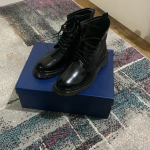 Svarta boots från Trussardi Jeans - Snygga svarta boots från Trussardi Jeans i glansigt skinn med grov sula och snörning framtill. Klassisk rund tå och robust design som passar perfekt till streetwear eller en mer dressad look. Kommer i originalkartong.