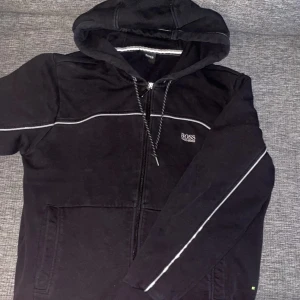 Svart hoodie från BOSS med dragkedja - Svart hoodie från BOSS med vit piping längs ärmar och bröst. Jackan har huva med dragsko, dragkedja framtill och två fickor. Perfekt för en avslappnad stil och enkel att matcha med det mesta.