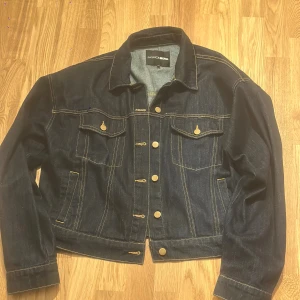 Mörkblå jeansjacka från Fashion Nova - Snygg mörkblå jeansjacka från Fashion Nova i storlek L. Jackan har klassisk design med guldfärgade knappar, två bröstfickor med lock och markerade sömmar. Tillverkad i 100% bomull för en skön och autentisk denimkänsla. Perfekt för dig som gillar streetstyle.