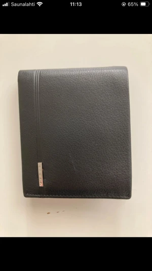 Svart plånbok i skinn från Cross - Stilren svart plånbok från Cross i äkta skinn med slät och lätt texturerad yta. Plånboken har flera fack för kort och ett myntfack med lock. Metallogo på framsidan ger en snygg detalj. Perfekt storlek för fickan och håller dina grejer organiserade.