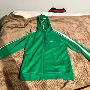 Grön set från BOSS med vita ränder - Snygg grön set från BOSS med vita ränder längs ärmarna och broderad logga på bröstet. Tröjan har huva med snörning, dragkedja framtill och två fickor. Perfekt för chill eller sportiga dagar, riktigt fräsch färg och klassisk streetstil. Använd fåtal gånger, jättebekväm tracksuit.