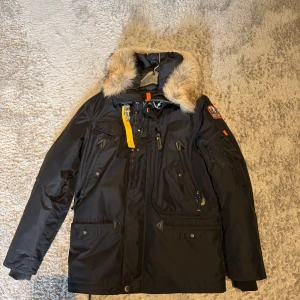 Svart Parajumpers parkajacka med päls - Svart parkajacka från Parajumpers med äkta päls på huvan och coola detaljer som gul rem och flera fickor med dragkedja. Jackan har lång ärm, hög krage och patch på axeln. Perfekt för kalla dagar och har en riktigt snygg streetstil.