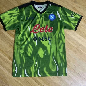 Napoli grön matchtröja EA7 - Säljer en Napoli fotbollströja från EA7 i grön med svart och vit detaljer. Tröjan har korta ärmar, V-ringad krage och ett unikt mönster som liknar eldflammor. Materialet är lätt och andas, perfekt för träning eller match. Tryck med Lete och MSC på bröstet.
