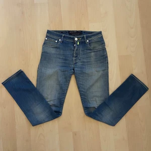 Jacob Cohën blå jeans - Snygga blå jeans från Jacob Cohën, modell 688.Comfort. Klassisk femficksdesign med vita knappar och gul kontrastsöm. Jeansen har en rak passform och är tillverkade i högkvalitativt denim med subtila slitningar för en cool look. Storlek W32. 